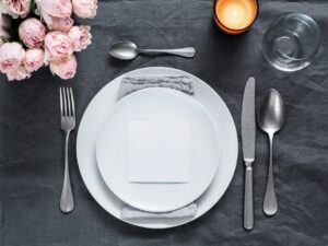 Looking down on a contemporary white place setting on a dark marble table with pink roses and silverware and glassware. 